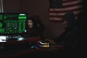 Two anonymous individuals wearing hacker masks use computers in a dark room with an American flag backdrop, highlighting cyber security themes.
